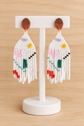 Boucles d’oreilles pendantes blanches en perles Miyuki avec motif géométrique multicolore, présentées sur un présentoir blanc et un fond bois clair
