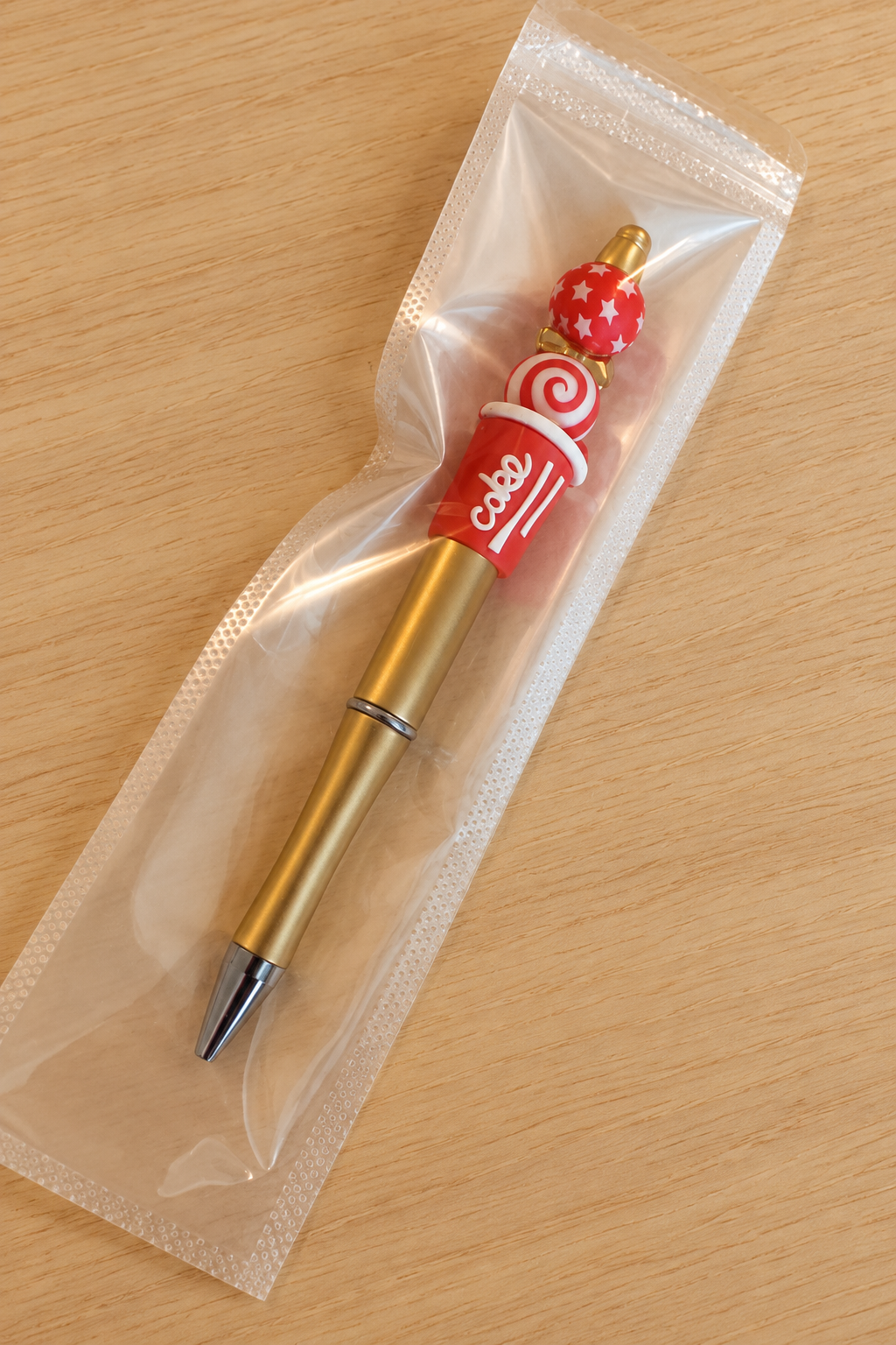 Stylo soda cola rouge et doré dans son sachet transparent sur bois clair