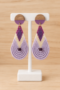 Boucles d’oreilles pendantes en perles violet, mauve et lilas présentées sur un présentoir blanc, sur fond bois clair
